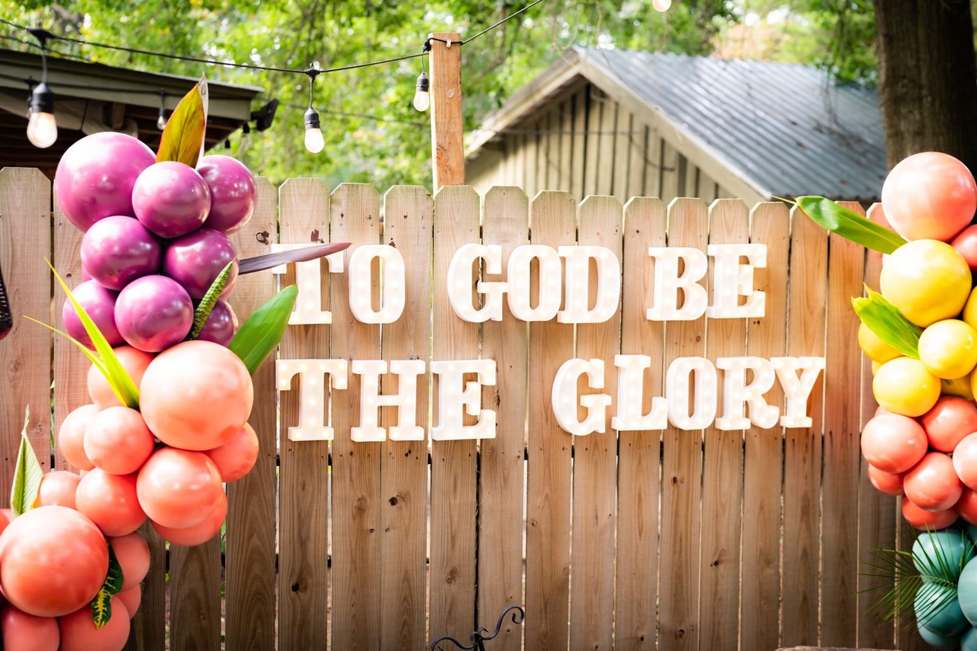 Marquee letters reading To God Be the Glory on a wooden fence, framed by magenta, coral, and yellow balloon clusters with tropical leaves under string lights