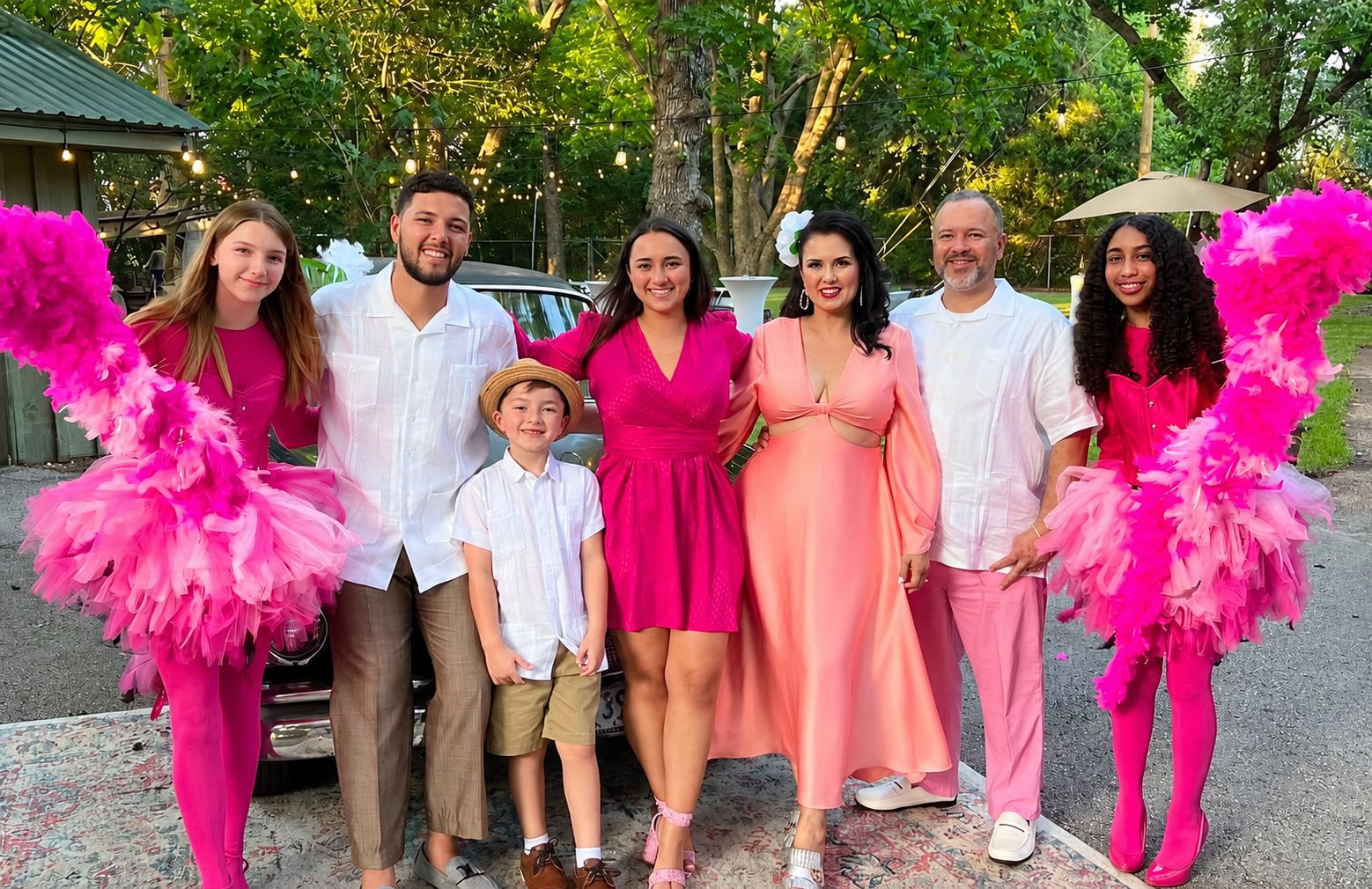 The Cuadra family dressed in pink and white guayaberas in front of a vintage car under string lights, with two dancers in pink feathered costumes.