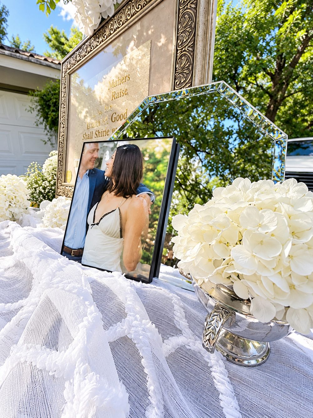 A welcome table with a white textured linen, an ornate gold framed sign, a framed photo of the couple, a beveled glass mirror, and white hydrangeas in a silver urn.