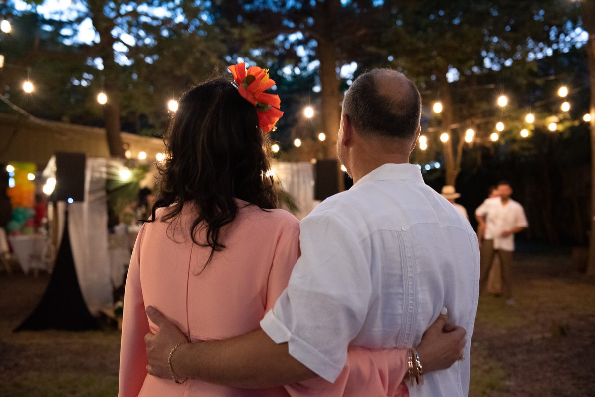 A couple in a coral dress and a white guayabera, arms around each other, watching the dance floor under string lights at a Havana Nights party.