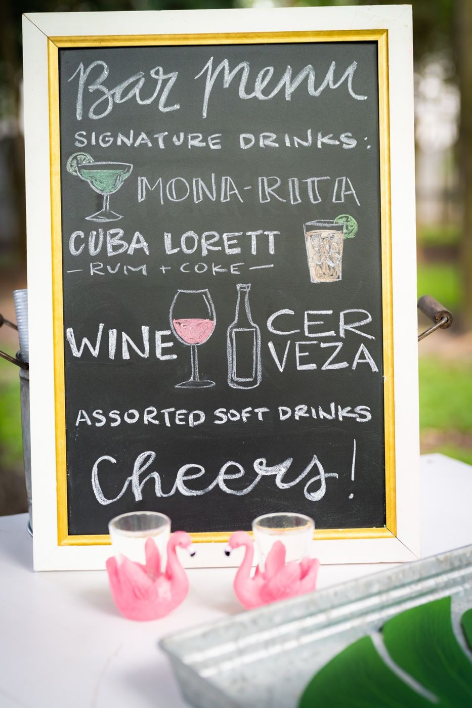 A chalkboard bar menu listing the Mona-Rita, the Cuba Lorett rum and coke, wine, and cerveza, with pink flamingo shot glasses in front