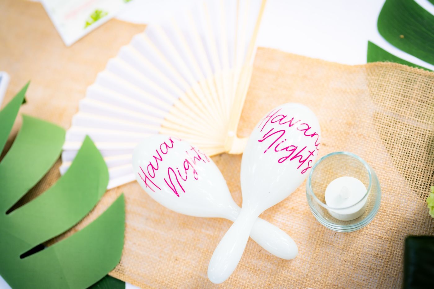 Two white maracas lettered Havana Nights in pink script beside a white paper fan, a votive, and monstera leaves on burlap