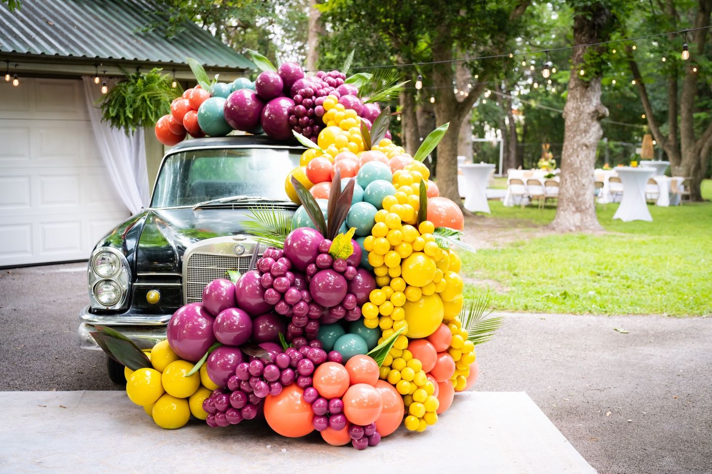 A vintage black Mercedes covered in a cascade of magenta, yellow, coral, and teal balloons with palm leaves, with string lights and cocktail tables behind it