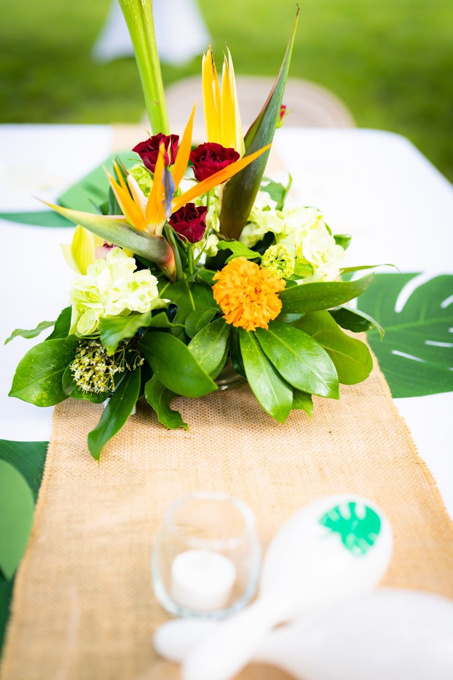 A tropical centerpiece of birds of paradise, red roses, green hydrangeas, and magnolia leaves on a burlap runner with monstera leaves
