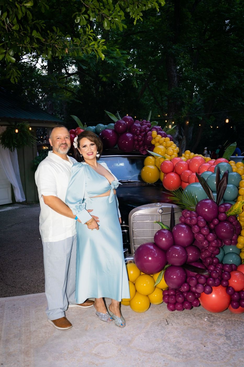 A smiling couple dressed in white and pale blue standing in front of the balloon-covered vintage car under the trees