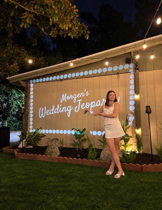 M.M. smiling beside her Wedding Jeopardy sign projected on the wall of The Magnolia Garden under string lights